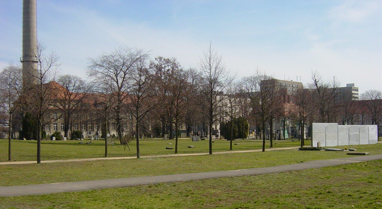 Broad lawn with scattered gravestones, trees and a surviving section of the Berlin Wall